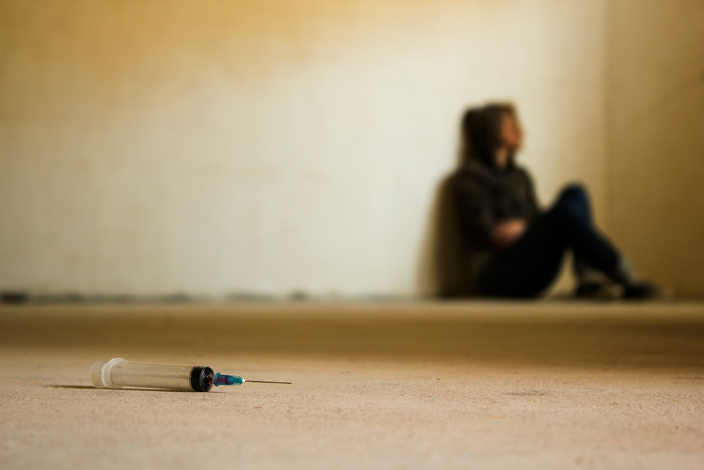 Addict sits in distance, needle visible in foreground